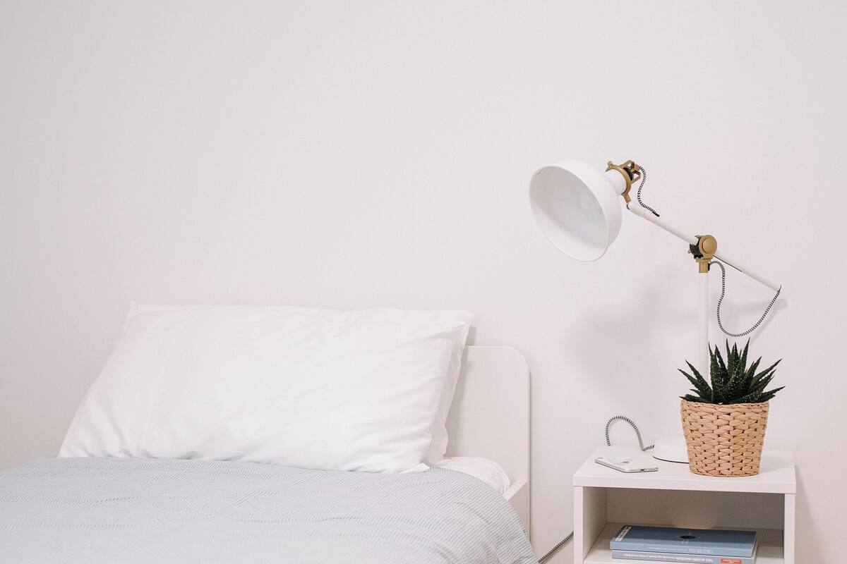 A neatly made bed with a white comforter and a white pillow. Next to the bed is a white nightstand with a white lamp, a woven basket with a plant, and a smartphone charging.