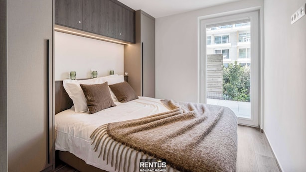 A modern bedroom with a large bed, wooden headboard, and a view of a building through a large window. - Port Orion - brand new ground floor flat with pool (Nieuwpoort)
