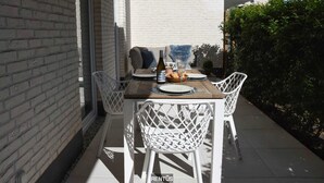 A patio with a table set for two, white chairs, and a bottle of wine. - Port Orion - brand new ground floor flat with pool (Nieuwpoort)