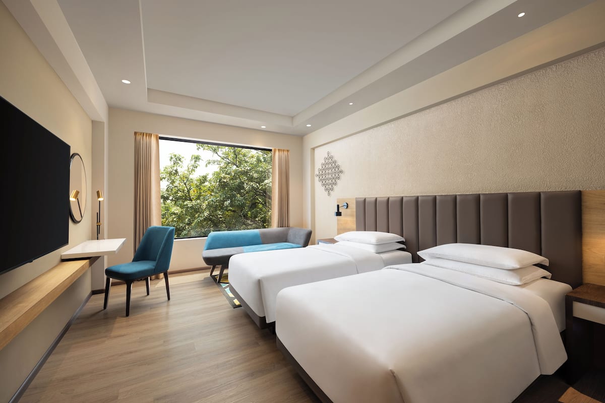 A hotel room with two beds, a desk, a chair, and a large window with a view of trees.