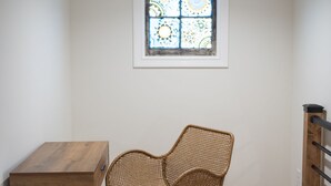 Una habitación de esquina con piso de madera, una silla de mimbre, una pequeña mesa de madera y un ventanal con diseño de vitral.