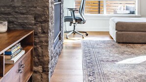 A modern living room with a stone fireplace, a flat-screen TV, a wooden bookshelf, a gray sofa, and a large window with blinds. - "Inspired Elegance" at Tamarack Lodge (Canmore)