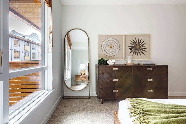 A bedroom with a large window, a wooden dresser, a mirror, and two framed artworks.