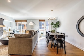 Uma sala de estar moderna com sofá, mesa de jantar e cadeiras. Há uma janela grande com persianas, um lustre e uma planta na parede.