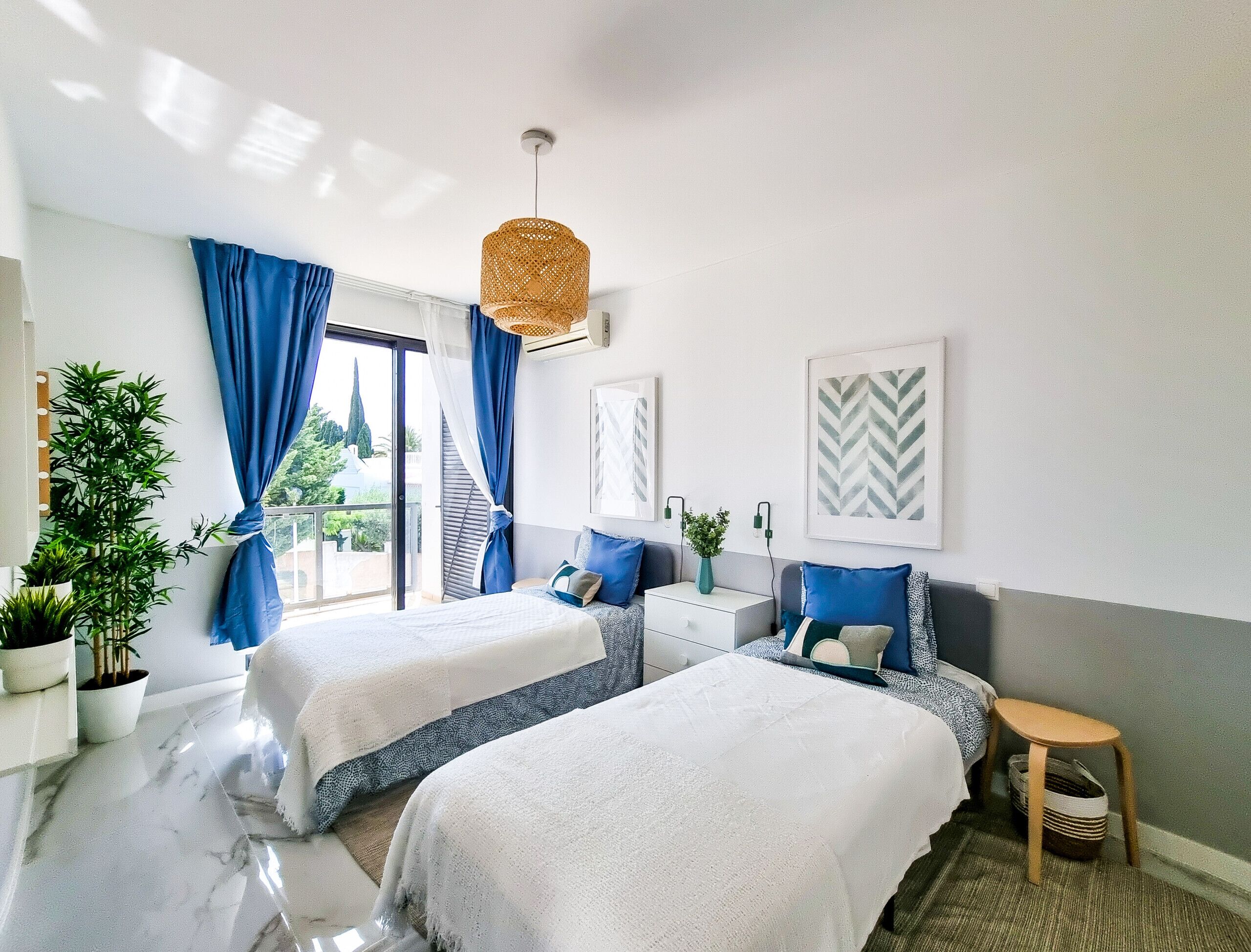 A hotel room with two beds, a large window with blue curtains, a hanging rattan light fixture, and a view of greenery outside.