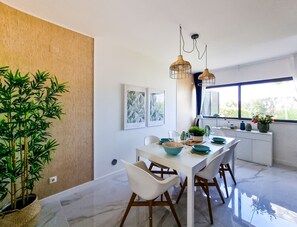 Uma área de jantar moderna com uma mesa branca, cadeiras e luminárias pendentes. Há quadros na parede, um vaso com planta e uma janela com vista para o verde lá fora.