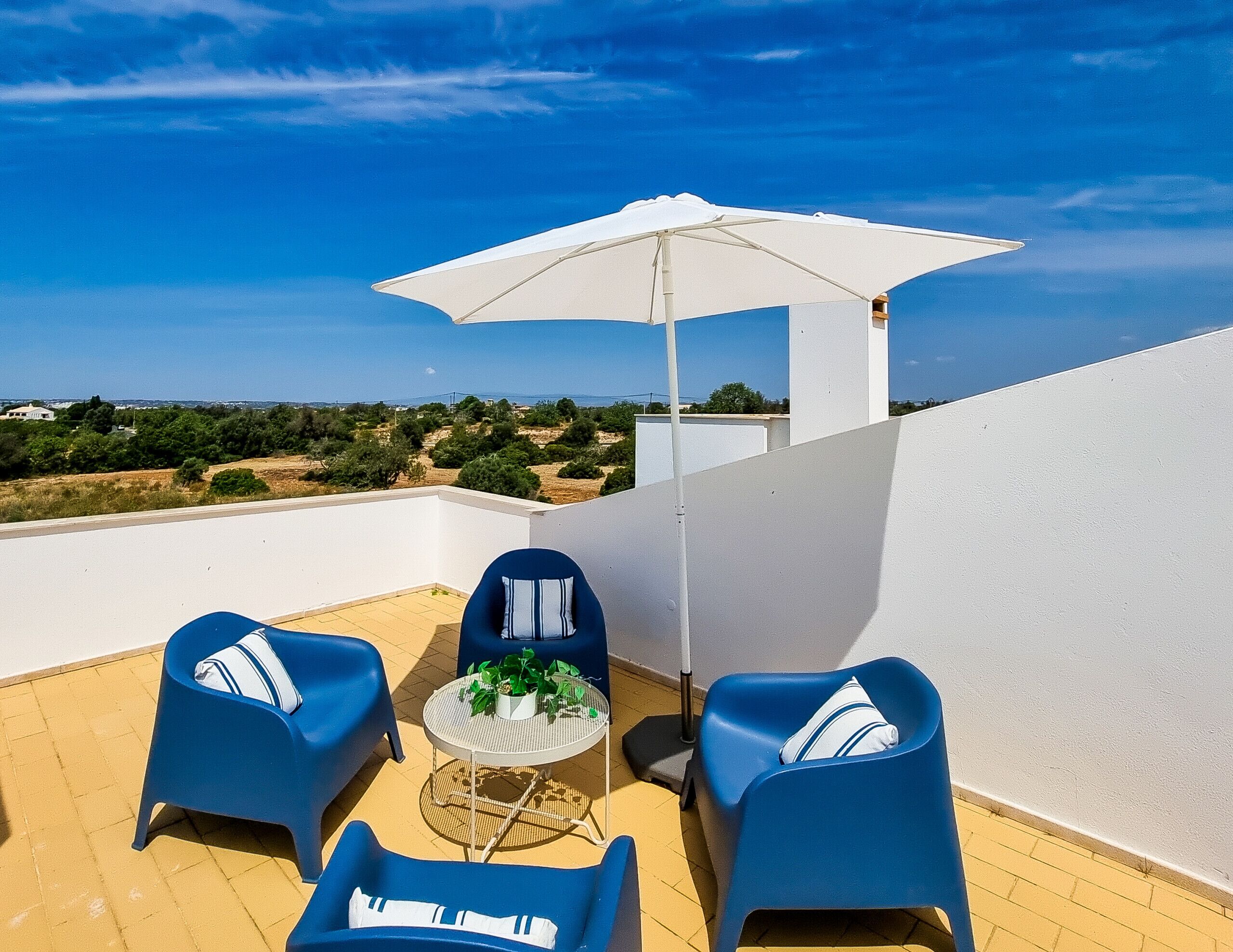 Una terraza en la azotea con dos sillas azules, una pequeña mesa redonda con una planta y un paraguas blanco.