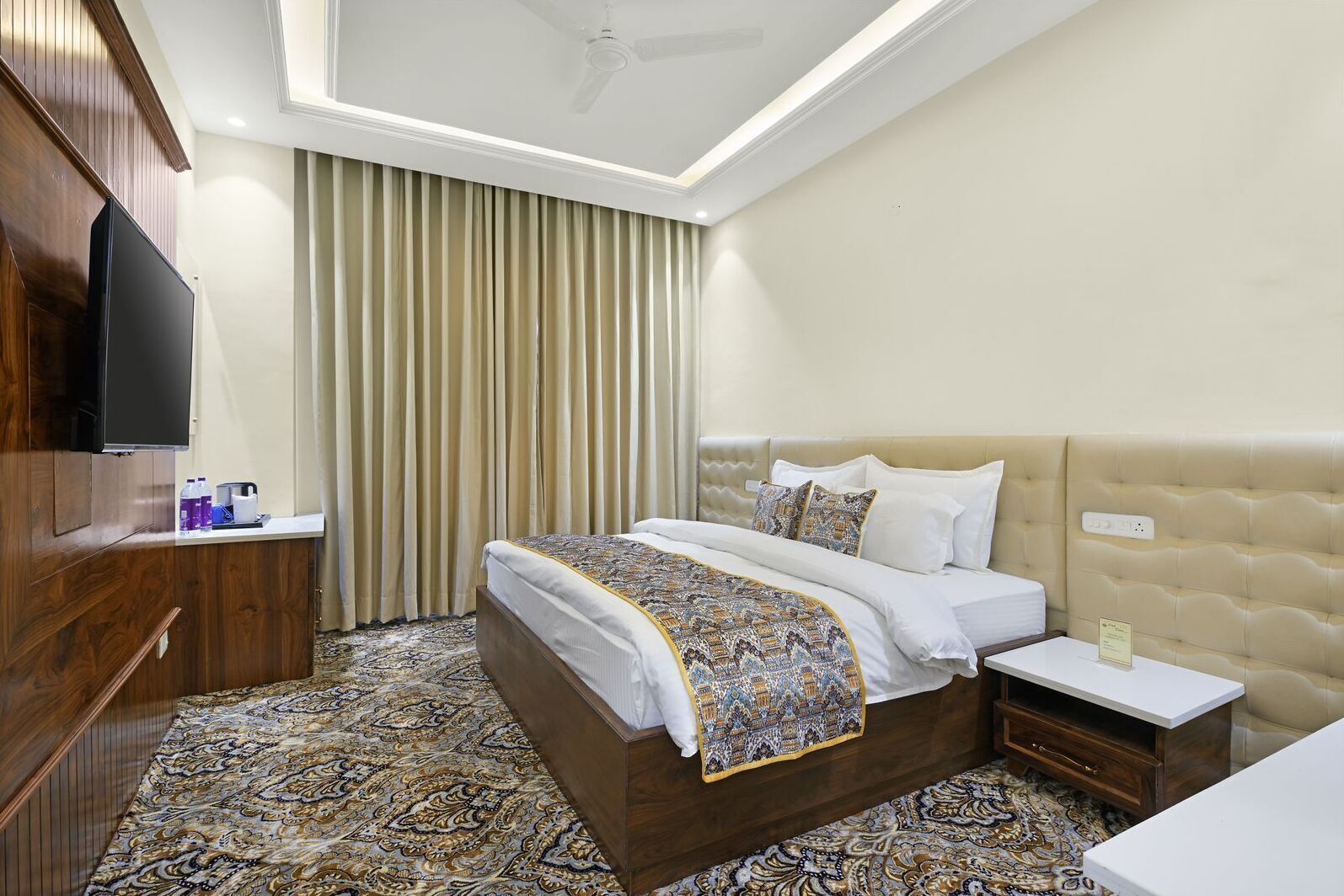 A hotel room with a large bed, a television on the left, a bedside table with a lamp, and a patterned carpet.