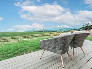 Two wicker chairs on a wooden deck with a view of green fields and distant hills.