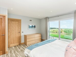 A bedroom with a large bed, a wooden dresser, a framed picture on the wall, and a view of a green landscape through a window.