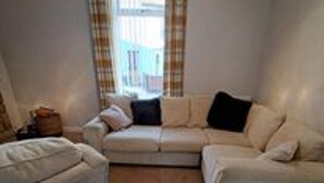 A living room with a beige sofa, a white armchair, and a window with curtains. - Immaculate 2-bed Cottage - Free Wifi (Bridlington)