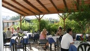 An outdoor dining area with tables covered in blue tablecloths, surrounded by greenery and a wooden pergola. - Gorgona Hotel (Malevizi)