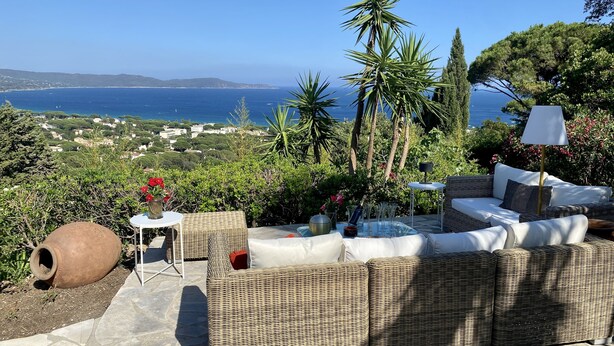 An outdoor seating area with wicker furniture, a table with a vase of flowers, and a view of the sea and distant landscape.