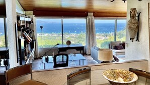 A spacious room with a large window offering a view of the sea and greenery. There is a dining area with a table and chairs, and a modern chandelier.