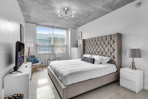 Relax in this serene bedroom with a large bed, TV, and a table with a lamp near the window offering a view of the outside.