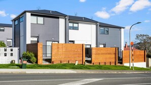 En moderne boligbygning i to etager med en facade i grå og hvid, et træhegn og en velholdt græsplæne.