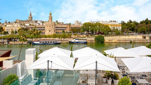 Une terrasse au bord de la rivière, avec des places assises en plein air et une vue sur des bâtiments historiques et des bateaux.