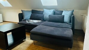 Avoin, loft-tyylinen huone, jossa on tummanharmaa kulmasohva, pieni musta pöytä ja kattovalokuvio.