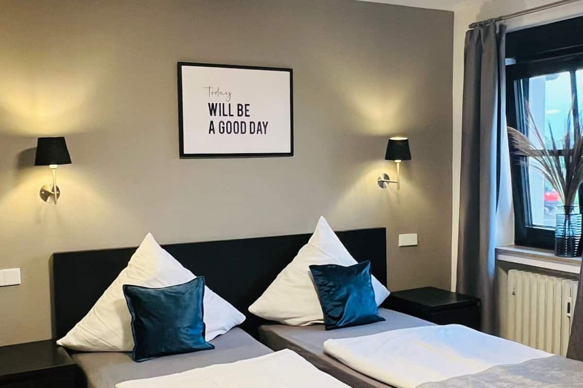 Quarto de hotel com duas camas, uma frase motivacional emoldurada na parede e uma janela com cortinas.