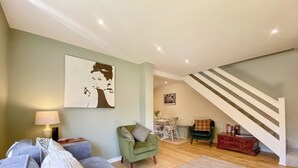 Una sala de estar con un sofá gris, un sillón verde, piso de madera, una escalera con barandillas blancas y un comedor con mesa y sillas.