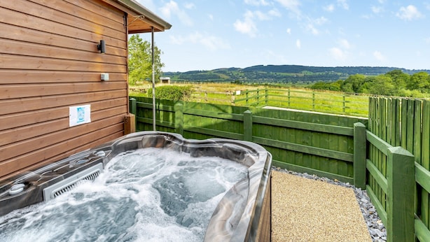 Jacuzzi exterior a construcción de madera con vista a un campo verde y colinas.