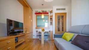 A modern living room with a flat-screen TV, wooden furniture, a dining table with chairs, and a sofa with cushions.