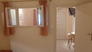 A room with a bed, a desk, and a chair. There is a window with curtains and a door leading to another room. - Aronis Studios Triple bed with View & Patio 03 (Kapsáli)