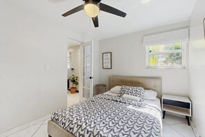 A bedroom with a bed, a bedside table, a ceiling fan, a window with curtains, and a door leading to another room.