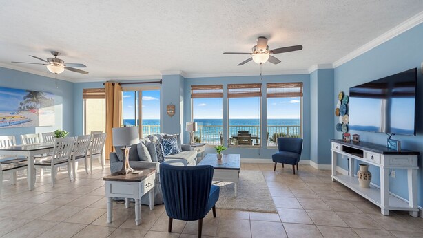 A spacious living room with a dining area, large windows with ocean views, ceiling fans, and a TV.