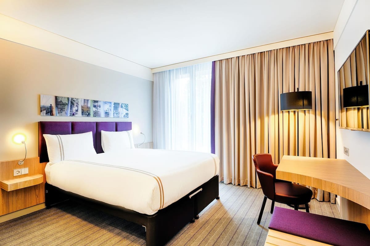 A hotel room with a bed, bedside table, lamp, chair, desk, and a large window with curtains.