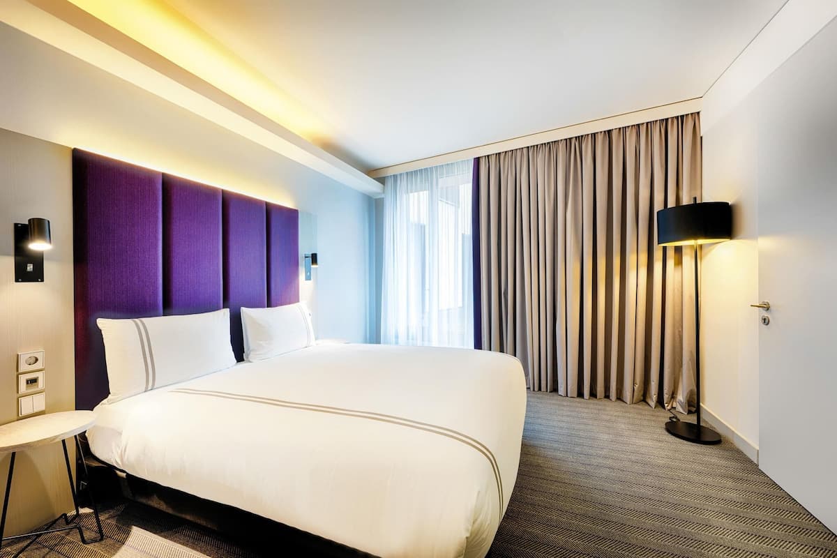 A hotel room with a large bed, a desk, a lamp, and a window with curtains.