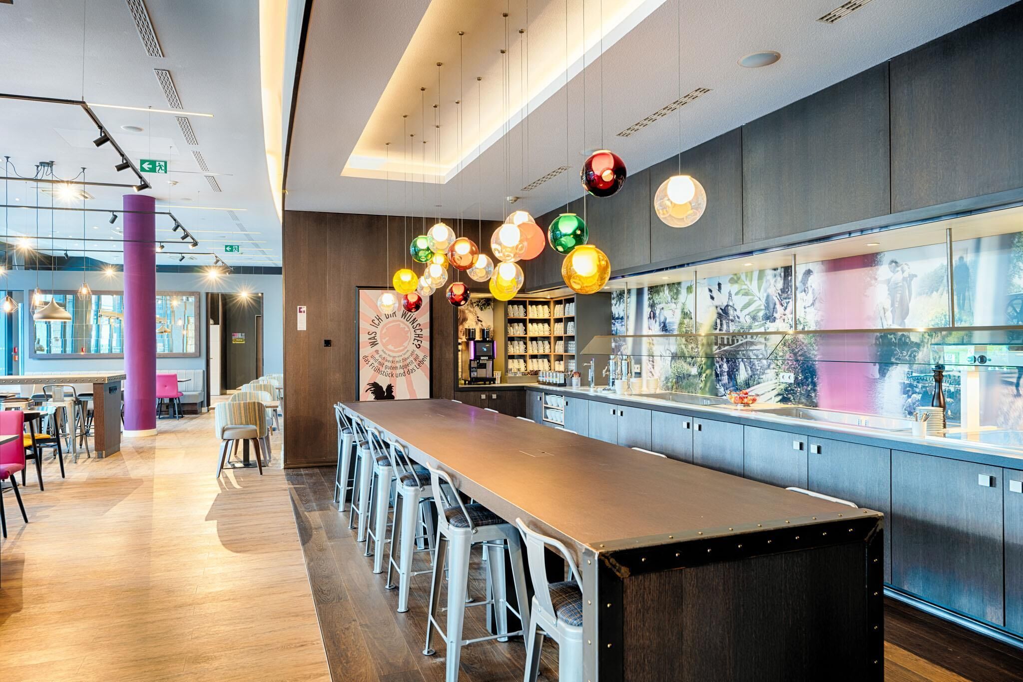A modern bar area with a long counter, bar stools, and colorful hanging lights.