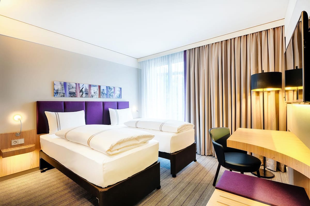 A hotel room with two beds, a desk, a chair, a nightstand, and a wall-mounted lamp.