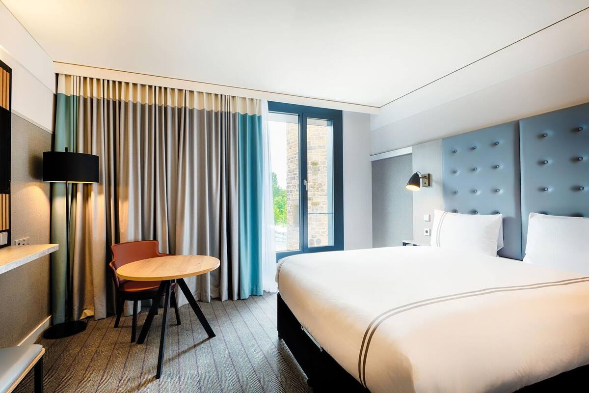 A hotel room with a bed, a chair, a table, a lamp, and a window with curtains.