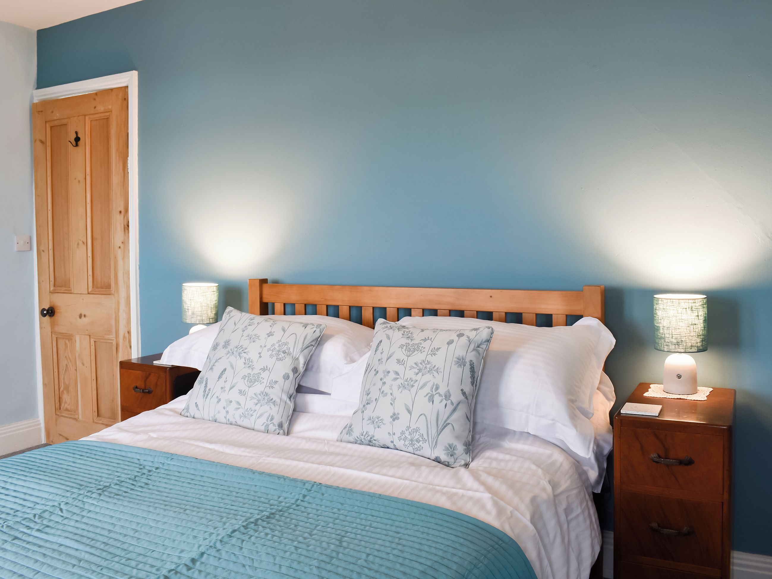 A bedroom with a wooden headboard, a blue wall, a wooden door, and two bedside tables with lamps.
