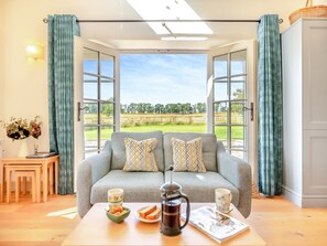 A cozy living room with a grey sofa, two patterned cushions, a coffee table with a coffee pot, a bowl of snacks, and a magazine. Large windows offer a view of a green field and trees. - 1 bedroom accommodation in Eglingham, near Alnwick (Eglingham, near Alnwick)