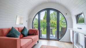 A cozy living room with a curved ceiling, a sofa with cushions, a large sliding glass door leading to a garden, and a small table with a remote control. - Woody's - 1 Bedroom Pod - Cold Inn (Kilgetty)