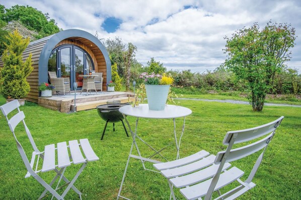 A modern outdoor seating area with a circular table and two white chairs, a small barbecue grill, and a wooden deck with outdoor furniture.
