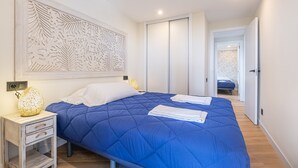 Un dormitori amb un llençol blau acolxat, un coixí blanc i un panell decoratiu a la paret. Hi ha una taula de nit amb una làmpada i un calaix petit.