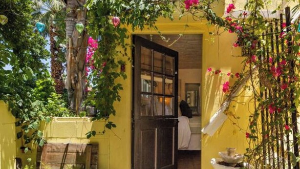 A yellow building with a dark door, potted plants, and hanging flowers. - Carrington Cottage @ 420 Shiloh LGBTA Pet friendly Sanctuary with Soul (Palm Springs)