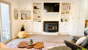 Una sala de estar con chimenea, muebles de cocina blancos, un televisor de pantalla plana y un sofá de cuero.
