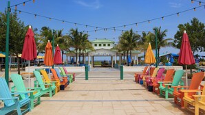 A beachside seating area with colorful chairs and umbrellas, a covered seating area in the background, and palm trees. - Private Beach Club, Heated Pool, Jacuzzi, & 6 Seater Golf Cart Included!  (Pompano Beach)