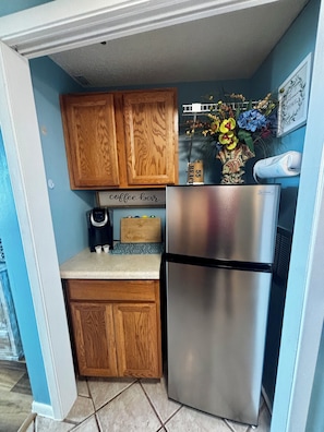 A compact kitchen with wooden cabinets, a stainless steel refrigerator, a coffee maker, and a decorative wall piece.