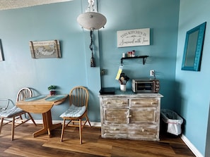 A kitchen with a wooden dining table, chairs, a hanging light fixture, a wall-mounted sign, a toaster, and a cabinet with drawers.