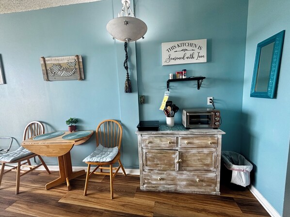 A kitchen with a wooden dining table, chairs, a hanging light fixture, a wall-mounted sign, a toaster, and a cabinet with drawers.