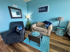 A living room with a dark armchair, a beige sofa, a coffee table, and a blue rug. There are two wall decorations, a framed mirror, and a sign that says 'Together is my favorite place to be.'