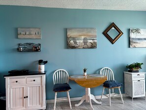 A room with a blue wall, a wooden table with two chairs, a white cabinet, and several wall-mounted paintings.