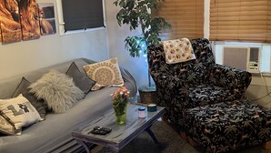 A living room with a grey sofa, a patterned armchair, a coffee table with a vase of flowers, a ceiling fan, and a large window with blinds. - The Lyons Den - Great work place with high speed internet near downtown.  (Jackson)