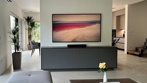 Ein modernes Wohnzimmer mit einem großen Flachbildfernseher, der an einem grauen Schrank befestigt ist, einem grauen Sofa, einem Couchtisch mit Blumenvase und einem Essbereich im Hintergrund.