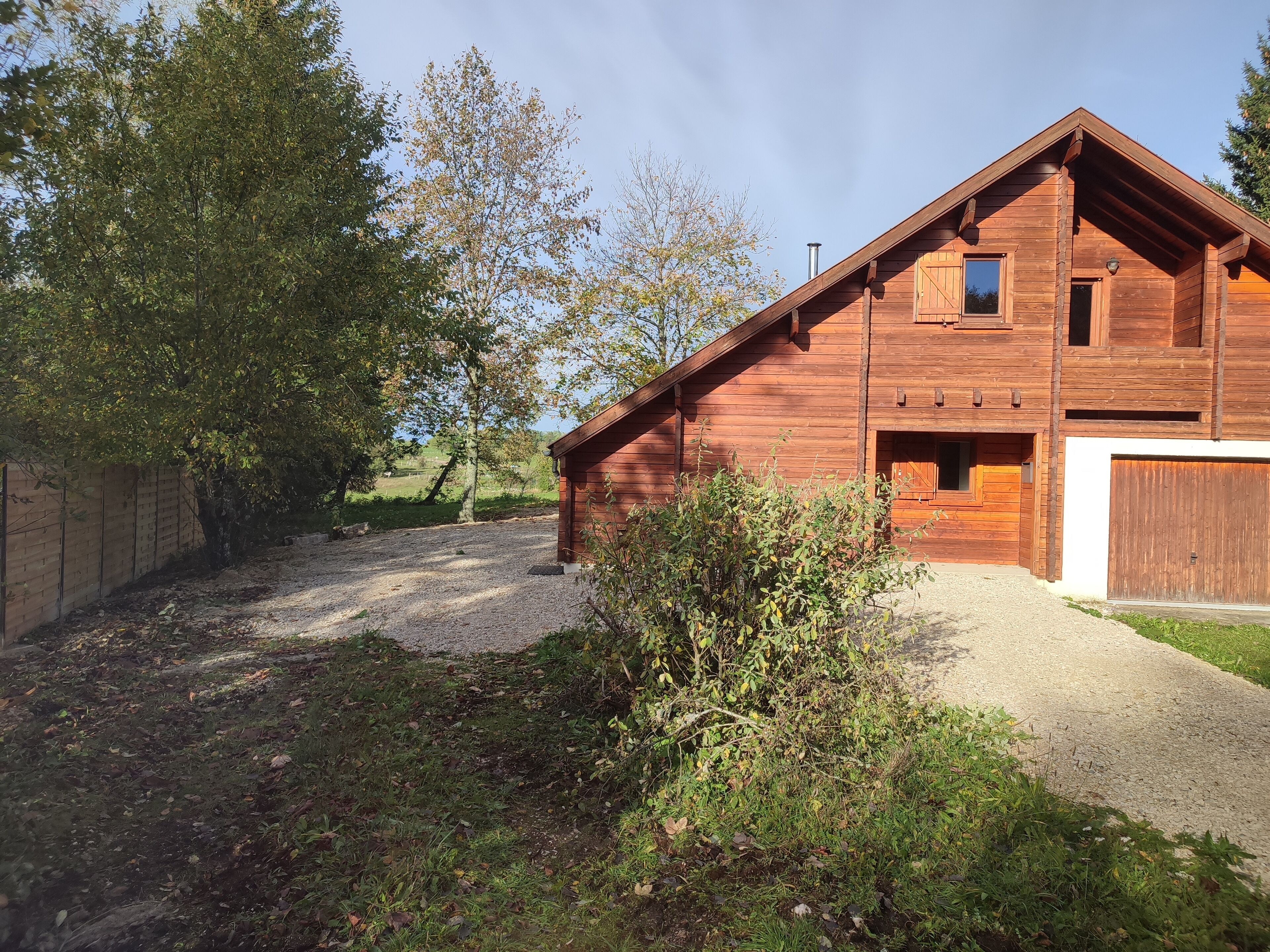 A wooden cabin with a gravel driveway, surrounded by trees and a fence.